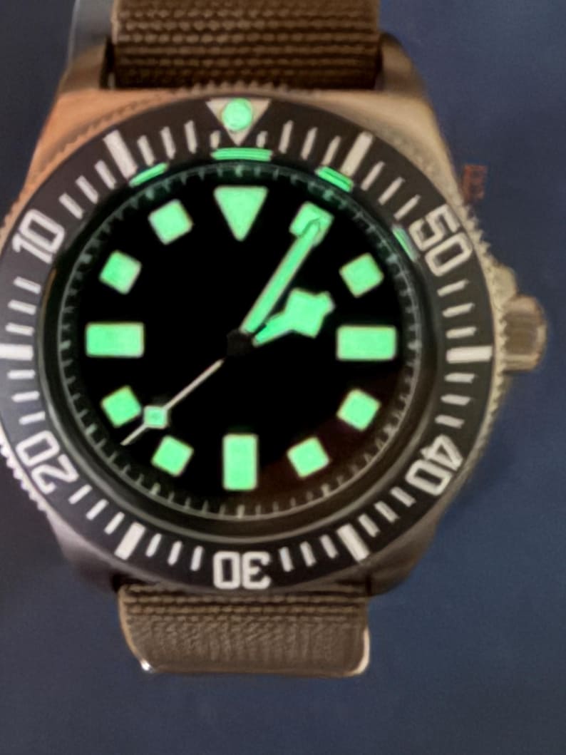 Peut inclure: Une montre-bracelet avec un cadran noir, des index lumineux verts et des aiguilles assorties. La montre pr&eacute;sente une lunette rotative noire avec des chiffres blancs et un bracelet en tissu marron. Le design inclut un marqueur triangulaire &agrave; douze heures et une trotteuse avec une petite pointe lumineuse carr&eacute;e.