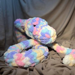 Può includere: Un morbido giocattolo a forma di serpente in peluche nei colori pastello rosa, giallo, blu e viola. Il serpente è fatto di un filato grosso e strutturato e ha un piccolo occhio nero. Il giocattolo è appoggiato su un tessuto color neutro.