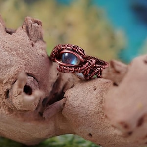 May include: A handcrafted copper wire ring featuring a blue-grey gemstone, intricately woven into a unique eye-shaped design. The ring is set against a natural wood background, showcasing the artisan's detailed wire-wrapping technique.