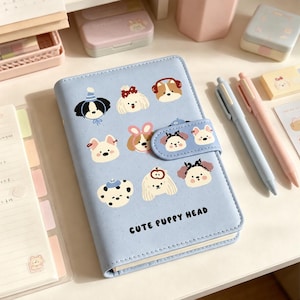 May include: A light blue notebook with a cute puppy head design. The notebook features various puppy illustrations and the text "CUTE PUPPY HEAD". Two pens, one light blue and one pink, are next to the notebook.
