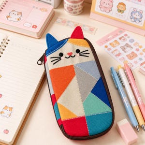 May include: A rectangular fabric pencil case shaped like a cat with a geometric patchwork design in red, orange, yellow, blue, teal, and gray. The case features embroidered black eyes, whiskers, a pink nose, and two upright ears with a dark brown zipper.