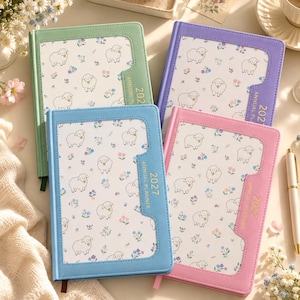 May include: Four 2027 annual planners with solid-colored covers in green, purple, blue, and pink. Each planner features a central white panel with a repeating pattern of small sheep and tiny flowers, and the text 2027 ANNUAL PLANNER.