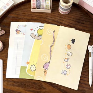 May include: Four rectangular sheets of stationery paper featuring various illustrations. One sheet has a lined grid and a small wizard cat, another shows a yellow cat in a UFO, a third displays a purple yarn ball, and the last features a vertical row of small cats.