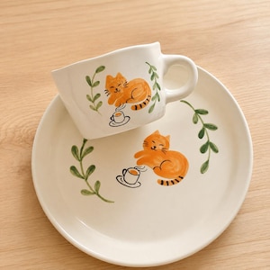 May include: A white ceramic cup and matching saucer set featuring hand-painted orange tabby cats. Each piece displays a cat sitting beside a small cup of coffee, framed by green leafy vines. The cup has a rounded handle and a smooth finish.