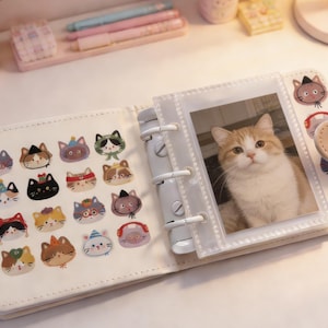May include: Open, cream-colored mini photo album with a photo of a cat in a clear sleeve. The album features cat-themed stickers and a beige strap with cat illustrations. The album is open on a white surface.