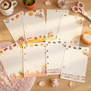 Puede incluir: Una colección de hojas de papel crema con líneas y pegatinas decorativas con temática de comida. Las pegatinas presentan ilustraciones de varios aperitivos y golosinas. También se ve una taza de chocolate caliente, una pequeña planta y otros artículos de papelería.