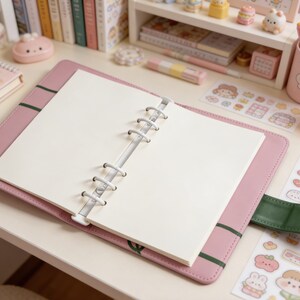May include: A pink faux leather binder-style notebook with a green accent. The notebook is open, revealing blank white pages. The desk is decorated with stickers, a mug with pens, and other stationery items. The overall theme is cute and organized.