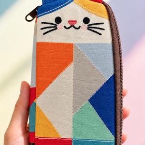 May include: A rectangular fabric pencil case featuring a cat face design with one blue ear and one red ear. The body is decorated with a geometric patchwork pattern in vibrant colors including orange, yellow, red, blue, teal, gray, and white.