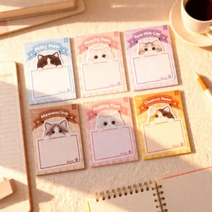 Può includere: Sei blocchi di note colorati a tema gatto. Ogni blocco note presenta un'illustrazione di gatto diversa con testo come "Milky Maw" e "Peachy Paws". I blocchi note hanno uno spazio vuoto per scrivere e un'impronta di zampa.