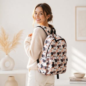 May include: A backpack with a repeating pattern of cartoon cats in various colors, including black, brown, and white. The backpack has black straps and a front pocket. The person is wearing a cream-colored sweater.