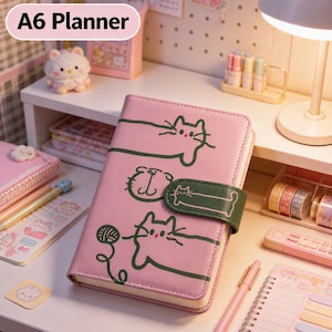 May include: Pink A6 planner with green cat illustrations and a green strap closure. The planner has the text "A6 Planner" in a white oval. Other stationery items are visible on the desk.