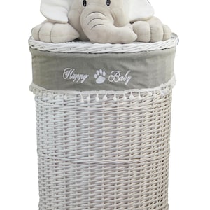 Puede incluir: Una cesta de lavandería de mimbre blanco con un forro de tela gris y un juguete de elefante de peluche. La tela tiene bordadas las palabras "Happy Baby". La cesta es cilíndrica y tiene un ribete decorativo.
