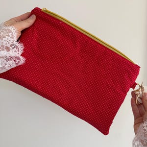 Peut inclure: Une pochette rouge avec une fermeture &eacute;clair dor&eacute;e et un petit motif de points r&eacute;p&eacute;t&eacute;s. La pochette est tenue par une personne portant une manche en dentelle blanche. Un porte-cl&eacute;s dor&eacute; est attach&eacute; &agrave; la pochette.