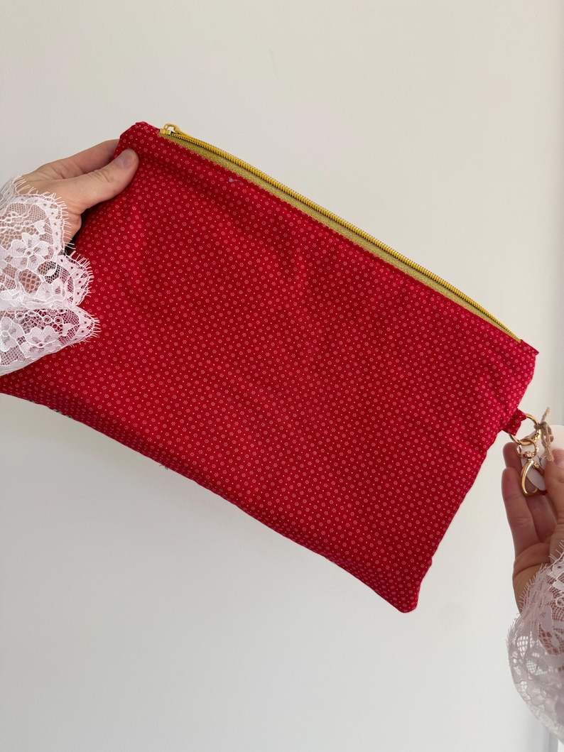 Peut inclure: Une pochette rouge avec une fermeture &eacute;clair dor&eacute;e et un petit motif de points r&eacute;p&eacute;t&eacute;s. La pochette est tenue par une personne portant une manche en dentelle blanche. Un porte-cl&eacute;s dor&eacute; est attach&eacute; &agrave; la pochette.