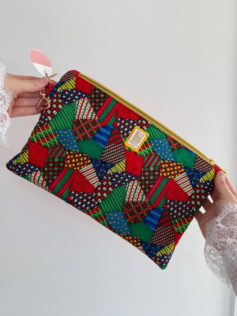 Peut inclure: Une pochette rectangulaire en patchwork color&eacute; avec une fermeture &eacute;clair dor&eacute;e. Le tissu pr&eacute;sente un m&eacute;lange vibrant de motifs g&eacute;om&eacute;triques rouges, verts, bleus et jaunes. Une petite &eacute;tiquette est suspendue &agrave; la tirette. La pochette est tenue par une personne.