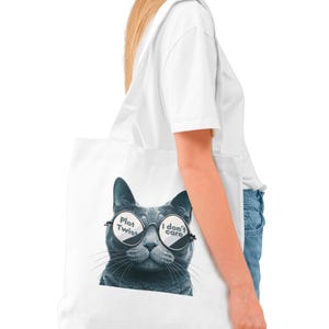 Pode incluir: Saco tote branco com um gato cinzento a usar óculos de sol. Os óculos de sol têm o texto "Plot Twist" e "I don't care". O saco é transportado ao ombro.