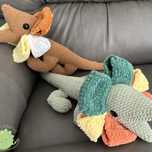 Peut inclure: Deux peluches dinosaures au crochet faites à la main, marron et vert. Le dinosaure marron a des accents orange, blancs et jaunes. Le dinosaure vert a des accents orange, jaunes et turquoise. Les deux ont des yeux en bouton noirs. Le logo "Stitch-a-saurus" est en bas à gauche.