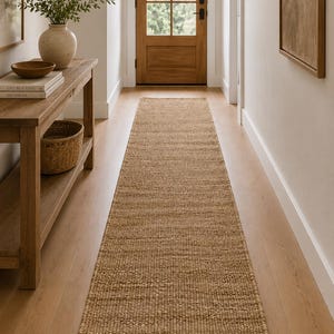 Pode incluir: Um tapete corredor retangular tecido em cor bege natural. O tapete está colocado sobre um chão de madeira clara num corredor, conduzindo a uma porta de madeira. A textura do tapete é visível, adicionando um elemento rústico ao espaço.