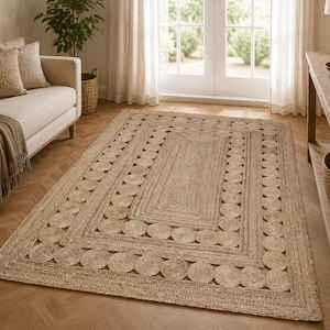 May include: A rectangular area rug crafted from natural tan jute fibers. The rug features a solid rectangular center panel surrounded by a decorative border composed of repeating circular woven jute motifs, creating a textured surface.