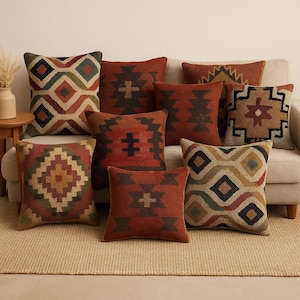 Puede incluir: Una colección de cojines decorativos con diseños geométricos en tonos rojos, marrones, verdes y beige. Los cojines están colocados sobre un sofá beige, creando un ambiente acogedor y atractivo. Los cojines varían en tamaño y forma.