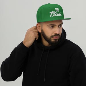Może przedstawiać: Zielona czapka z daszkiem z napisem "GO Birds" w białym skrypcie. Czapka ma strukturalny krój i płaski daszek. Czapka jest noszona przez osobę noszącą czarną bluzę z kapturem.