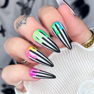 Puede incluir: Diseño de uñas stiletto con rayas blancas y negras y detalles de pintura goteando en verde neón, azul, amarillo y rosa. Las uñas largas y puntiagudas crean una declaración audaz y artística.