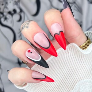 Puede incluir: Primer plano de uñas largas y puntiagudas con un diseño rojo, negro y blanco. Algunas uñas presentan una forma de corazón rojo, mientras que otras tienen una base negra con un acento blanco y rojo. Las uñas están bien cuidadas y llamativas.