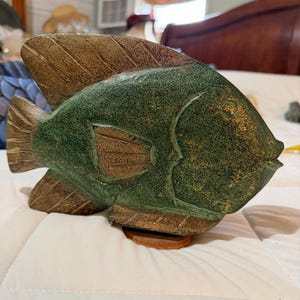 May include: A hand-carved wooden fish sculpture with a green body and brown fins. The fish is resting on a small, round, brown wooden base. The sculpture has a textured appearance and is likely a decorative item.