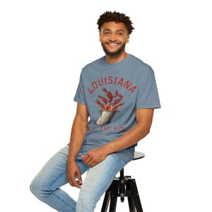 May include: A light blue t-shirt with the word "LOUISIANA" in red, above a graphic of crawfish boiling in a pot. The text "HOME OF THE BOIL" is below the pot. The shirt is worn by a person.