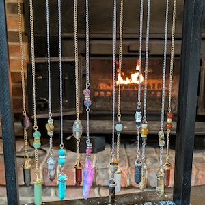 May include: A display of pendulums hanging from chains. The pendulums feature various crystals and beads in shades of green, blue, purple, and clear. The display stand is black with a decorative design at the base. A fireplace is visible in the background.