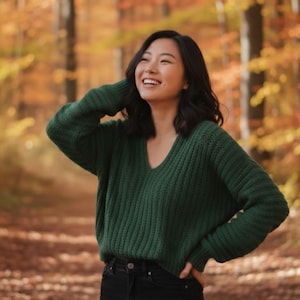 May include: A woman wearing a dark green V-neck sweater and black jeans smiles outdoors. The sweater has a ribbed knit texture. The background features a blurred view of a forest with autumn foliage.