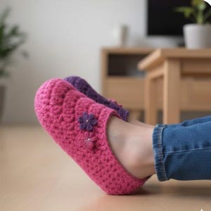 Könnte beinhalten: Handgefertigte, pinkfarbene Häkelschuhe mit lila Blumenakzenten. Die Schuhe werden von einer Person getragen, die eine blaue Jeans trägt. Diese gemütlichen Schuhe sind für den Innenbereich konzipiert.