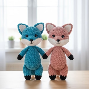 Könnte beinhalten: Zwei handgefertigte Fuchs-Stofftiere aus Häkelarbeit. Einer ist türkisblau mit weißen und schwarzen Akzenten, der andere ist rosa mit weißen und schwarzen Akzenten. Beide haben ein lächelndes Gesicht und stehen aufrecht.
