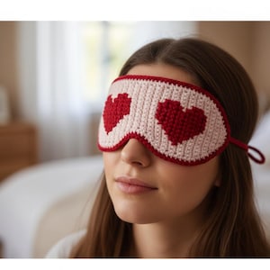 May include: A cream-coloured, crocheted sleep mask featuring two red hearts. The mask has a red border and ties. The image shows a close-up of the mask on a person's face, suggesting a focus on sleep and relaxation.