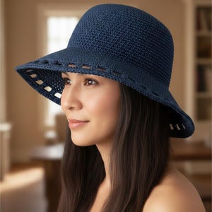 Könnte beinhalten: Ein marineblauer Häkel-Sonnenhut mit breiter Krempe. Der Rand hat ein dekoratives Ausschnittmuster. Der Hut hat eine runde Krone und besteht aus einem strukturierten, gewebten Material. Der Hut ist zum Sonnenschutz konzipiert.