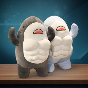 May include: Two plush shark-shaped toys. One is grey and the other is light blue. Both have white bellies with a muscle design, small arms, and open mouths with red interiors. They stand on a wooden surface against a blue background.