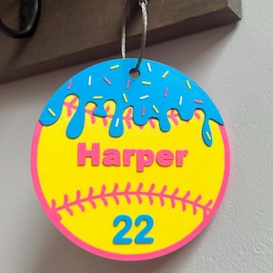 May include: A softball-themed keychain with a yellow base, pink trim, and blue icing detail. The name "Harper" is in pink, and the number "22" is in blue. The keychain is hanging from a metal loop.