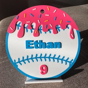 May include: A baseball-themed decoration with a donut design. The top features pink frosting with colorful sprinkles. The name "Ethan" is in blue letters, and the number "9" is in pink. The baseball has blue stitching and a blue border.