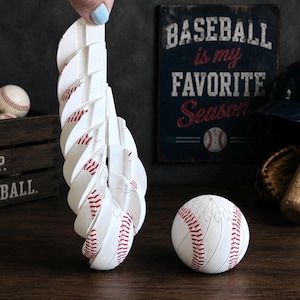 May include: A white baseball-themed sculpture featuring a series of stacked, curved segments that mimic the appearance of a baseball with red stitching, accompanied by a single, spherical baseball-patterned object resting on the surface.