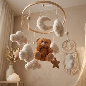 Può includere: Mobile per bambini in legno con un orsacchiotto marrone, soffici nuvole bianche, stelle e una luna crescente. Il mobile è sospeso a un anello di legno. La palette di colori è neutra con toni morbidi e caldi.