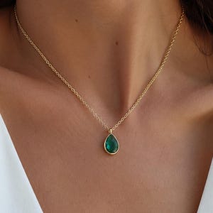 Può includere: Una collana a catena dorata con un ciondolo a forma di goccia caratterizzato da una pietra verde scuro sfaccettata incastonata in una cornice dorata. La collana poggia sulla clavicola, mostrando un design a maglie sottili e finitura metallica.