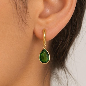 May include: A gold hoop earring featuring a dangling, faceted, pear-shaped green gemstone. The earring is secured through the earlobe, showcasing a polished gold metal finish and a vibrant, deep green translucent stone with a smooth, teardrop-shaped cut.