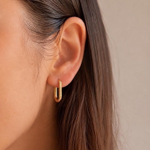 Könnte beinhalten: Goldfarbene, rechteckige Creolen. Die Ohrringe haben ein schlichtes, elegantes Design mit poliertem Finish. Das Bild zeigt eine Nahaufnahme des Ohrs und der Ohrringe.