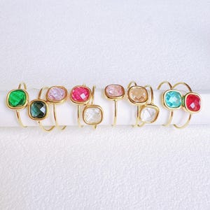 Puede incluir: Una colección de anillos de metal dorado, cada uno con una piedra preciosa de corte cuadrado en varios colores. Las piedras preciosas incluyen verde, turquesa, rosa, transparente y melocotón. Los anillos se exhiben sobre una superficie blanca.