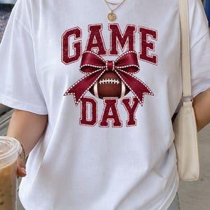 Woman wearing a white Comfort Colors Game Day football shirt with a maroon bow graphic at a football stadium.