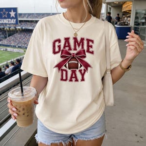 Woman wearing an Ivory Comfort Colors football Game Day shirt with a maroon coquette bow graphic.