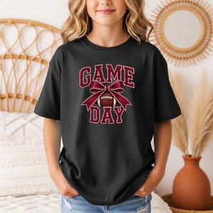 Youth black Comfort Colors football Game Day shirt with a maroon coquette bow design.