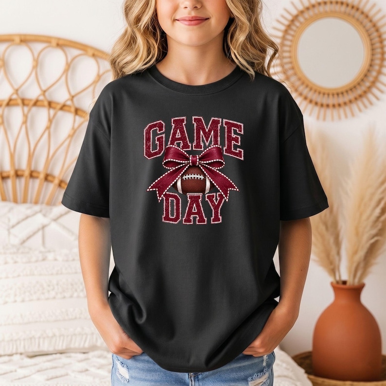 Youth black Comfort Colors football Game Day shirt with a maroon coquette bow design.