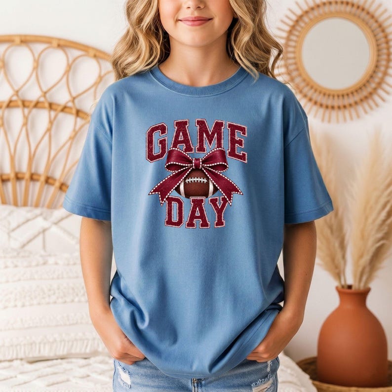 Youth Blue Jean Comfort Colors Game Day football shirt featuring a maroon bow graphic.