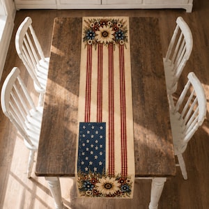 Puede incluir: Un camino de mesa rectangular con un diseño patriótico que presenta rayas de cuadros vichy en rojo y blanco, un campo azul con estrellas blancas y detalles florales que incluyen girasoles y pequeñas flores azules y rojas en los extremos.